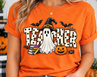 May include: An orange short-sleeve t-shirt featuring a graphic print with the word TEACHER in white letters with black polka dots. The design includes a white ghost wearing a black witch hat, a stack of colorful books, a pencil, a jack-o'-lantern, bats, and a spider.