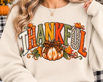 May include: A cream-colored crewneck sweatshirt featuring the word THANKFUL in large, multi-patterned letters. The design includes a central orange pumpkin with a plaid bow, surrounded by autumn leaves, berries, and vines in fall colors.