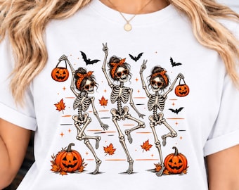 May include: A white t-shirt featuring a graphic print of three skeletons with messy buns, orange polka-dot headbands, and heart-shaped sunglasses. The skeletons are dancing among orange autumn leaves, black bats, and two jack-o'-lantern pumpkins.