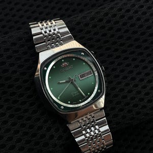 May include: A silver-tone Orient wristwatch with a green dial, featuring a day-date window at the three o'clock position, silver hands, and a metal link bracelet. The dial displays the Orient logo and the text Crystal 21 Jewels.