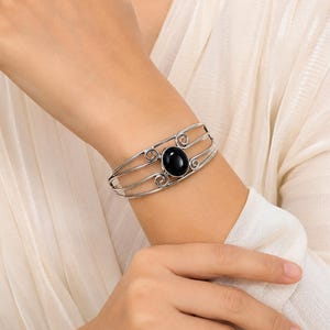 Könnte beinhalten: Ein silberfarbenes Armband mit einem zentralen ovalen schwarzen Stein. Das Armband besteht aus drei parallelen Metalldrähten, die sich an den Verbindungspunkten zur Steinfassung zu dekorativen Spiralen formen.
