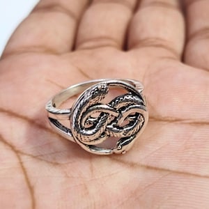 May include: A silver-toned metal ring featuring a circular design composed of two intertwined snakes. The metal has an oxidised finish that highlights the textured scales and detailed heads of the snakes, forming a continuous loop band.
