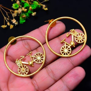 May include: A pair of gold-toned circular earrings featuring a bicycle silhouette centred within each hoop. The earrings have a matte finish and include a post-back closure mechanism. The bicycle design is detailed with spokes on the wheels.