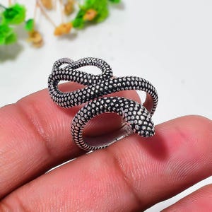 Può includere: Un anello in metallo color argento a forma di serpente avvolto. La superficie dell'anello presenta un motivo testurizzato simile a squame lungo tutta la sua lunghezza, con la testa del serpente posizionata a un'estremità del design aperto.