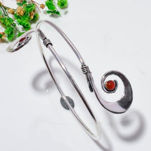 May include: A silver-toned metal arm cuff featuring a bypass design with two spiraling ends. Each end is accented with a small, round, polished orange-red carnelian gemstone. The metal band has a smooth, reflective finish with wire-wrapped details.