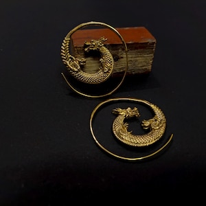 Può includere: Un paio di orecchini a spirale color oro con un motivo a drago dettagliato. Ogni orecchino è realizzato in forma circolare con il corpo del drago che forma la curva, mostrando squame testurizzate e una testa definita con corna.