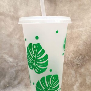 May include: A frosted translucent plastic tumbler with a white lid and a matching straw. The cup features a repeating pattern of vibrant green monstera leaves and small solid green dots scattered across the exterior surface of the container.