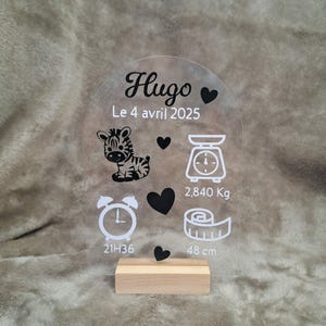Peut inclure: Une plaque en acrylique transparent en forme d'arche sur un socle en bois avec des motifs imprimés noirs et blancs incluant le nom Hugo, la date 4 avril 2025, un zèbre, une balance avec 2,840 Kg, une horloge à 21H36 et un mètre ruban de 48 cm.