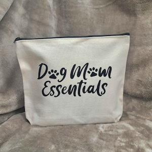 Peut inclure: Une pochette zippée en toile couleur crème avec le texte noir "Dog Mom Essentials" imprimé sur le devant. La lettre "o" dans "Dog" et "Mom" est remplacée par un motif de patte noire. La pochette possède une fermeture éclair noire.