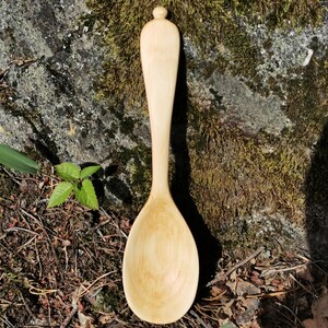Könnte beinhalten: Ein heller Holzlöffel mit einer glatten, ovalen Laffe und einem langen, schlanken Stiel, der oben in einem kleinen, abgerundeten Knauf endet. Die Holzmaserung ist auf der gesamten Oberfläche sichtbar und zeigt natürliche Variationen in hellen Beigetönen.