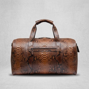 Könnte beinhalten: Eine braun-schwarze Reisetasche mit texturiertem Schlangenhautmuster. Die Tasche verfügt über zwei Tragegriffe, eine horizontale Reißverschlusstasche auf der Vorderseite und einen Hauptreißverschluss. Das Design zeigt ein wiederkehrendes Schuppenmuster.