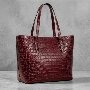 Op de afbeelding: Een diepbordeauxrode tote bag met een gestructureerd krokodillenpatroon. De tas heeft een gestructureerde rechthoekige vorm met twee verticale handvatten aan het bovenpaneel, wat zorgt voor een consistente donkerrode afwerking over de buitenkant.
