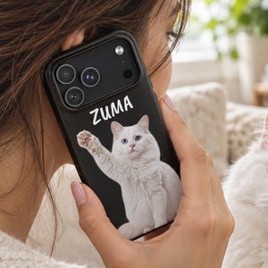 Pode incluir: Uma capa de smartphone preta com um gato branco de olhos azuis e o nome ZUMA impresso em letras brancas acima do gato. O gato é retratado com uma pata levantada e a capa tem um recorte para um sistema de câmara de lente tripla.