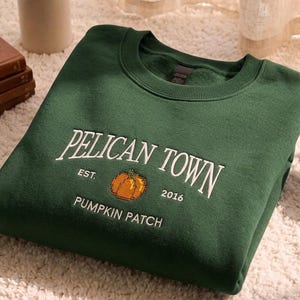 May include: A dark green crewneck sweatshirt featuring white embroidered text that reads PELICAN TOWN, EST. 2016, and PUMPKIN PATCH, with a small orange pumpkin graphic centered between the text. The garment has a ribbed collar and cuffs.