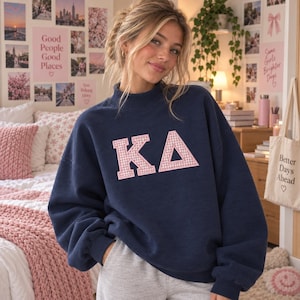 May include: A navy blue long-sleeve sweatshirt featuring a mock neck collar and the Greek letters KA in a pink and white gingham pattern on the chest. The sweatshirt has a relaxed fit with ribbed cuffs and a ribbed hem, constructed from fabric.