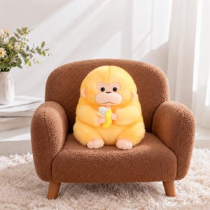 May include: A plush yellow monkey toy with a light-colored face and ears, holding a small yellow and white banana. The monkey is sitting on a brown, textured, upholstered armchair with short wooden legs and rounded armrests.