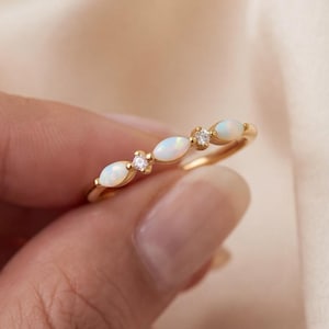 Può includere: Un anello in oro con tre opali taglio marquise e due piccole pietre rotonde trasparenti disposte in fila lungo la fascia. Le pietre si alternano tra le forme di opale e pietra trasparente, creando un design simmetrico sulla fascia.