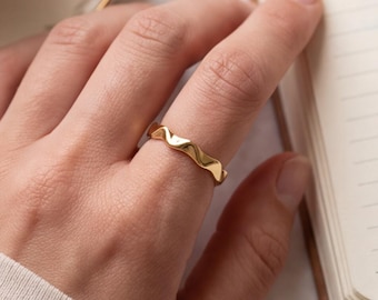 May include: A gold-toned ring with a continuous wavy, undulating band design. The ring features a polished, reflective metallic surface that catches the light, creating a series of smooth, rhythmic curves around the circumference of the finger.