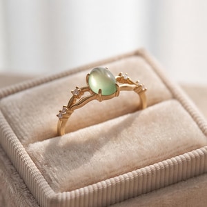 May include: A gold ring featuring a central oval-shaped light green cabochon gemstone held by four prongs. The band is crafted with a delicate, branching design that includes two small, clear, faceted stones set on each side of the central gem.