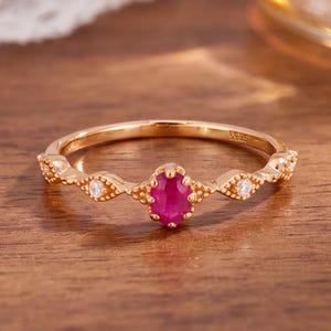May include: A rose gold-toned ring featuring a central oval-cut vibrant pinkish-red gemstone held in a prong setting. The band is accented with two small round clear stones on either side, set within decorative metal frames with beaded details.