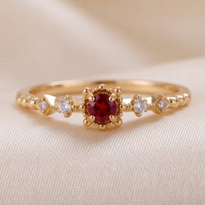 May include: A gold ring featuring a central round-cut red gemstone held by a beaded setting. The band is accented with two smaller round white stones and two diamond-shaped white stones, all set in gold with a textured, beaded metal finish.