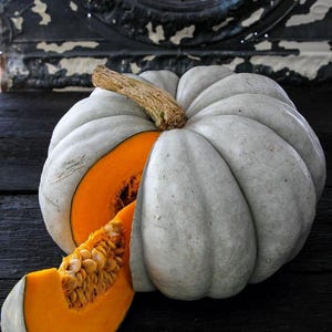 May include: A large, round, light grey pumpkin with deep vertical ridges and a thick, textured brown stem. A wedge-shaped slice has been removed from the side, revealing the bright orange interior flesh and a cluster of light tan pumpkin seeds.