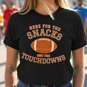 Puede incluir: Una camiseta negra de manga corta con un gráfico de un balón de fútbol americano en el centro. El texto "HERE FOR THE SNACKS AND THE TOUCHDOWNS" está impreso en letras mayúsculas de color naranja sobre y bajo la ilustración del balón.