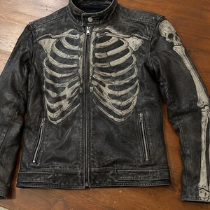 May include: A black leather jacket featuring a distressed finish and a prominent off-white skeleton rib cage graphic printed across the front. The left sleeve displays a matching skull and arm bone print. It includes a stand-up collar, a central zipper closure, and two zippered side pockets.
