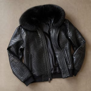 Pode incluir: Um blusão bomber preto com um padrão texturizado de relevo de crocodilo em todo o exterior. O casaco inclui um colarinho de pelo preto espesso e felpudo, um fecho de correr central e punhos de malha canelada preta nas mangas e na bainha.