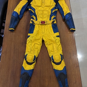 Può includere: Una tuta integrale gialla e blu con dettagli modellati simili a un'armatura sul petto, sulle spalle e sulle ginocchia. Il costume include una cintura abbinata con un emblema centrale rosso a forma di X, maniche lunghe e pannelli integrati.