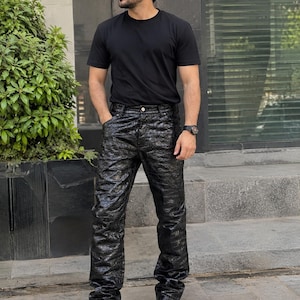 May include: A black short-sleeved crew neck t-shirt paired with black trousers featuring a glossy, textured crocodile-skin pattern. The trousers have a button and zip fly closure, and the outfit is completed with white athletic trainers.