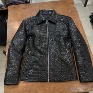 Könnte beinhalten: Eine schwarze Jacke mit texturiertem Krokodilprägemuster, ausgestattet mit einem spitzen Kragen, langen Ärmeln und einem durchgehenden silberfarbenen Metallreißverschluss in der Mitte. Zwei diagonale Reißverschlusstaschen vorne.