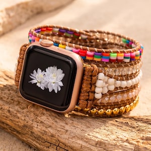 May include: A rose gold smartwatch with a black screen displaying two white flowers, attached to a multi-strand wrap bracelet featuring brown cord, colorful flat beads, clear faceted beads, white round beads, and metallic gold spheres.