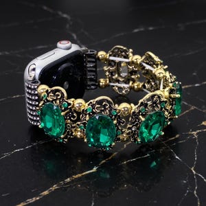 May include: A silver-toned smartwatch case attached to a decorative watch band featuring oval-cut emerald green gemstones set in ornate gold-tone metal frames, accented with small clear crystals and gold beads on a flexible elastic cord structure.