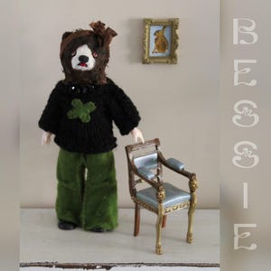 Puede incluir: Una muñeca con cabeza de oso, suéter negro con un trébol verde y pantalones verdes. La muñeca está junto a una silla en miniatura con asiento azul y detalles dorados. Una pequeña imagen enmarcada cuelga de la pared.
