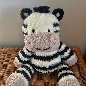 May include: A plush zebra toy crafted from fuzzy white, black, and light brown yarn. The stuffed animal features a white face with black stripes, a light brown muzzle, black ears, and a tuft of black hair on top of its head. It has a round body.