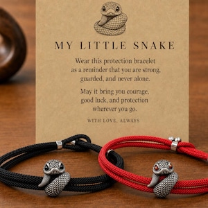 Könnte beinhalten: Zwei Kordelarmbänder, eines in Schwarz und eines in Rot, jeweils mit einem silberfarbenen Schlangenanhänger. Die Armbänder liegen vor einer beigen Karte mit einer Schlangenillustration und dem Text: MY LITTLE SNAKE. Wear this protection bracelet as a reminder that you are strong, guarded, and never alone. May it bring you courage, good luck, and protection wherever you go. WITH LOVE, ALWAYS.