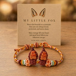Peut inclure: Un bracelet en cordon tressé avec deux petites breloques texturées en forme d'oreilles de renard dans des tons orange et marron. Le bracelet est présenté devant une carte beige portant le texte MY LITTLE FOX et un message sur l'amour.