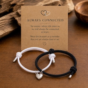 May include: A pair of adjustable cord bracelets, one white and one black, featuring a silver-toned magnetic heart charm. The bracelets are displayed on a rectangular kraft paper card with the text "ALWAYS CONNECTED" and a printed message.