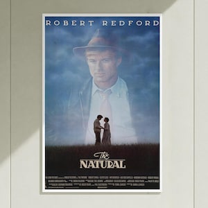 Peut inclure: Une affiche de film pour The Natural présentant un grand portrait estompé de Robert Redford portant un chapeau fedora au-dessus d'une scène plus petite de deux personnages debout dans un champ. Le titre The Natural est écrit en blanc en bas.