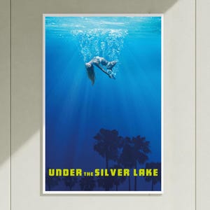 Puede incluir: Un póster rectangular vertical que muestra una escena submarina de color azul profundo con una figura nadando hacia abajo. Las siluetas de palmeras son visibles en la parte inferior contra el agua oscura. El texto UNDER THE SILVER LAKE aparece en amarillo.