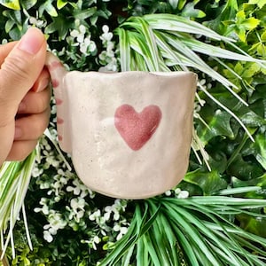 Może przedstawiać: Jasnoróżowy kubek ceramiczny o teksturowanej, nakrapianej powierzchni i nierównym brzegu. Z przodu kubka znajduje się pojedyncze, wyśrodkowane, ręcznie malowane ciemnoróżowe serce. Uchwyt ozdobiony jest naprzemiennymi poziomymi paskami w kolorze jasnego i ciemnego różu.