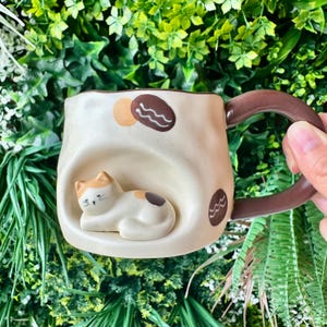 May include: A cream-colored ceramic mug with a speckled finish, featuring a three-dimensional white and orange cat resting in a recessed alcove. The mug has a dark brown handle and is decorated with small brown and orange abstract shapes.
