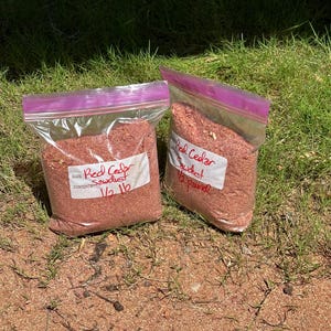 May include: Two clear plastic resealable bags filled with reddish-brown red cedar sawdust. Each bag features a white adhesive label with the handwritten text Red Cedar Sawdust 1/2 lb. The bags are positioned upright on a patch of green grass and dirt.