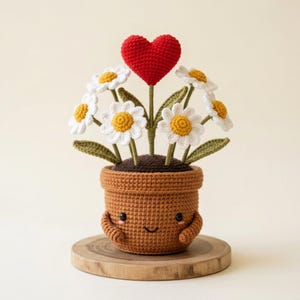 Può includere: Un vaso marrone all'uncinetto con un viso sorridente, piccole braccia e guance rosa, contenente un bouquet di margherite bianche con centri gialli e foglie verdi, sormontato da un cuore rosso, il tutto realizzato con filato lavorato a maglia.