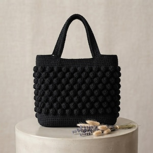 May include: A black crochet handbag featuring a textured bobble stitch pattern throughout the main body, a flat base, and two sturdy top handles. The bag is constructed from yarn with a consistent dark black color and a uniform, dense stitch.