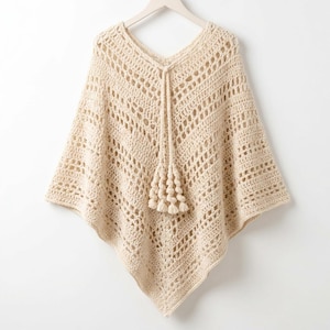 Op de afbeelding: Een crèmekleurige gehaakte poncho met een opengewerkt ruitpatroon en een V-hals. Het kledingstuk is voorzien van een centraal trekkoord dat eindigt in een bundel van vier decoratieve kwastjes die in het midden aan de voorzijde hangen.