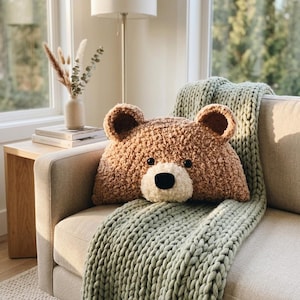 May include: A plush, brown, textured bear-shaped pillow with rounded ears, black button eyes, and a cream-colored muzzle featuring a black nose. The pillow is resting on a sofa draped with a thick, sage green, chunky knit throw blanket.