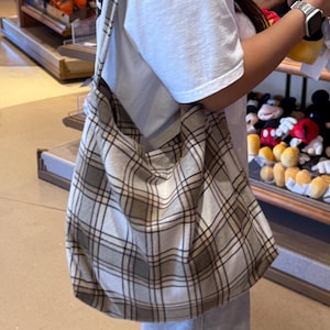 Peut inclure: Un sac à bandoulière de style hobo avec un motif à carreaux dans des tons beige, marron et blanc. Le sac possède une large bandoulière intégrée et une structure souple et affaissée, porté sur un t-shirt blanc uni et un pantalon de jogging gris.