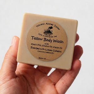 May include: A square bar of beige tallow body wash with a round brown paper label. The label features a volcano icon and text: Volcanic Azores Tallow, 100% Azores Grass-fed, Tallow Body Wash with Goat's Milk, Coconut Oil, Castor Oil, Scented with Litsea Cubeba.